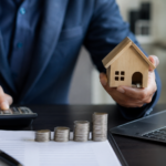 Profissional utiliza calculadora enquanto segura uma miniatura de casa, com moedas empilhadas e laptop sobre a mesa em operação de administração de garantias.
