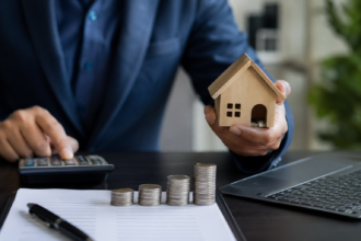 Profissional utiliza calculadora enquanto segura uma miniatura de casa, com moedas empilhadas e laptop sobre a mesa em operação de administração de garantias.