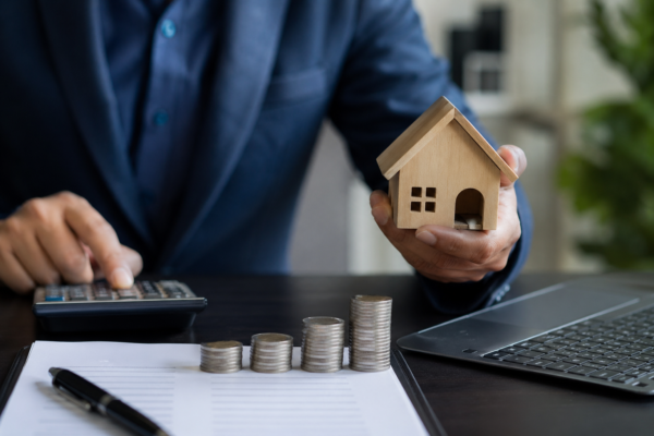 Profissional utiliza calculadora enquanto segura uma miniatura de casa, com moedas empilhadas e laptop sobre a mesa em operação de administração de garantias.